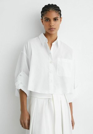 Femeie cu părul împletit, purtând o cămașă albă lejeră cu nasturi și mâneci suflecate, și o fustă plisată albă asortată, stând în fața unui perete alb.