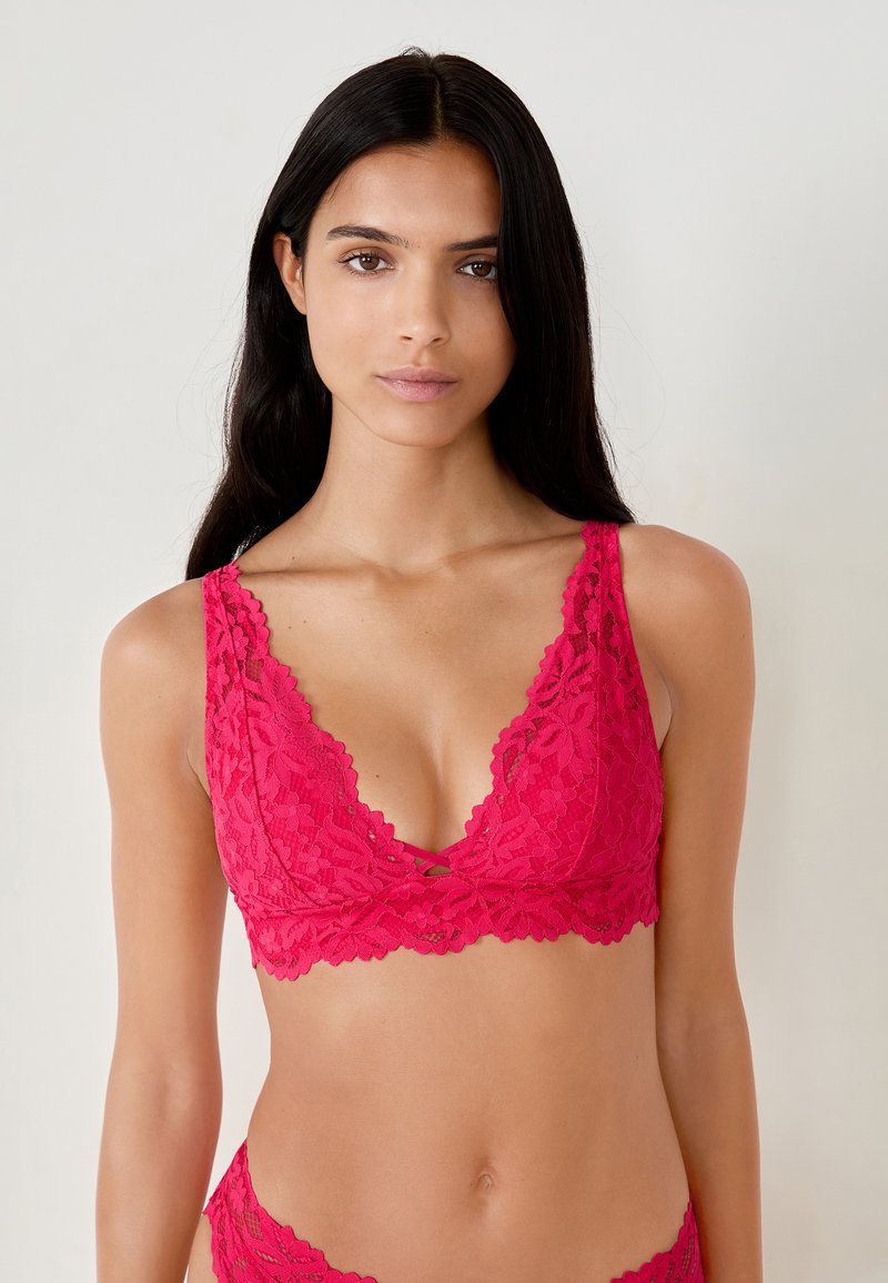 Bralette in pizzo di un vivace rosa con scollo a V profondo, bordi ondulati e motivi floreali. Presenta spalline regolabili e una texture morbida.