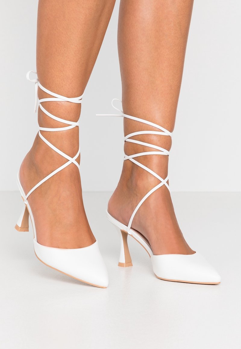 Pieds portant des escarpins blancs à bout pointu avec de fines brides enroulées autour des chevilles sur un fond clair uni.