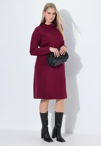 Abito lungo bordeaux a maniche lunghe con colletto alto, abbinato a una pochette nera e stivali neri con tacco al ginocchio. Tessuto dalla texture liscia.