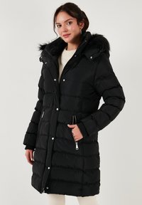 Chaqueta de plumas negra con capucha, que presenta un borde de piel sintética, bolsillos con cremallera y cierre de botones a presión. Texturizada con acolchado horizontal.