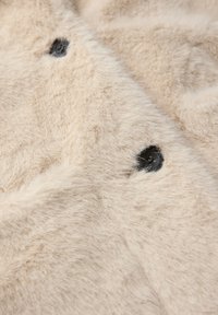 Tissu en fausse fourrure beige présentant une texture douce, avec deux boutons foncés visibles à la surface. Un éclat subtil ajoute de la profondeur.