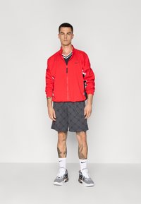 Rød zip-up jakke med hvite detaljer, svart t-skjorte, grå shorts med quiltemønster, og grå joggesko med svarte elementer.