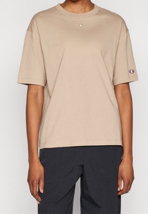 Persoon draagt een beige Champion T-shirt met korte mouwen en logo op de borst en mouw, gecombineerd met een zwarte broek.