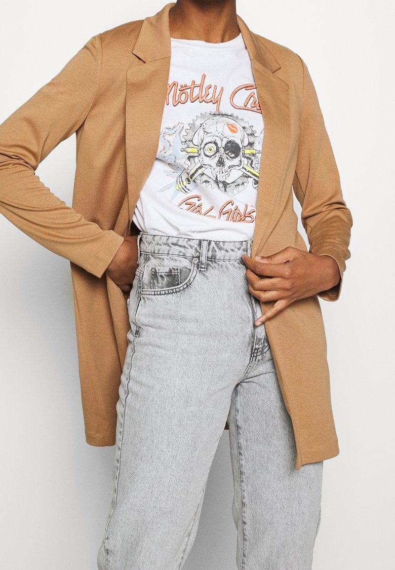 Personne portant un blazer marron clair, un t-shirt blanc à motif crâne et un jean gris clair taille haute, ajustant le devant du blazer avec la main.