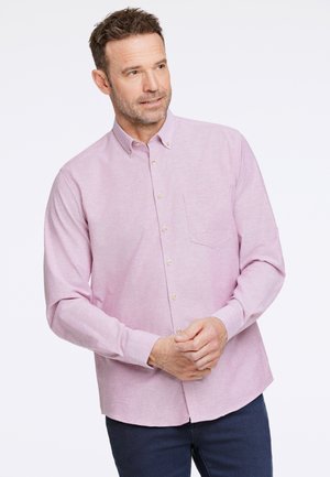 Camisa de botones rosa claro hecha de una tela texturizada, con un bolsillo en el pecho y un dobladillo curvado, combinada con pantalones vaqueros oscuros.
