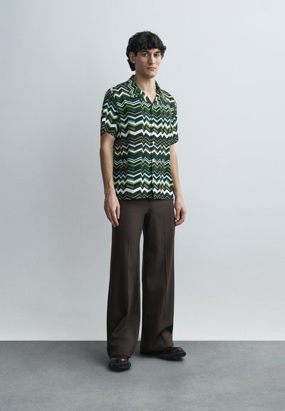 Κοντομάνικο πουκάμισο με πράσινο και μαύρο ζιγκ-ζαγκ μοτίβο, συνδυασμένο με φαρδιά καφέ παντελόνια και χοντρά μαύρα παπούτσια.