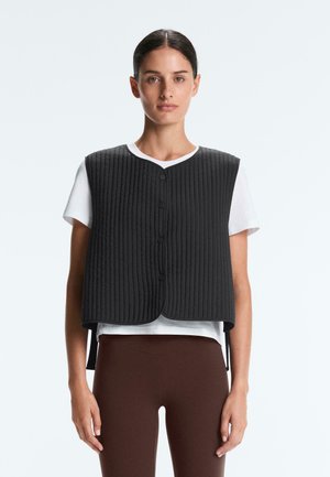 Žena v čiernej prešívanej vesti bez rukávov cez biele tričko, v hnedých priliehavých nohaviciach, stojaca pred jednofarebným svetlým pozadím.