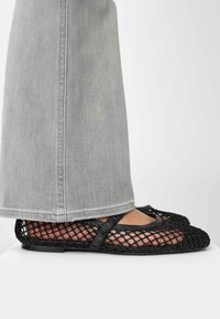 Zwarte mesh balletschoenen met een ronde neus en kruislings banden, gecombineerd met lichtgrijze wijde broekspijpen. De textuur is open en ademend.