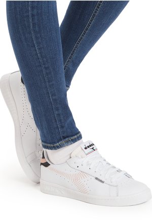 Sneaker bianche in pelle texturizzata, con perforazioni e accenti rosa/nera. Indossate con jeans azzurri aderenti, che mostrano un look casual.