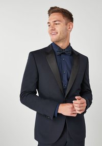 Giacca da smoking blu navy con rever in raso, abbinata a un papillon coordinato e una camicia azzurra attillata. Il tessuto appare liscio con un design strutturato.