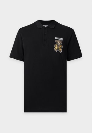 Νέος άνδρας που φοράει μαύρο πόλο Moschino με μπαλώματα αρκουδάκι, μαύρο παντελόνι με χρυσό φερμουάρ και μαύρες μυτερές μπότες.