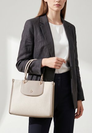 Femme en blazer sombre et top blanc tenant un grand sac à main beige texturé avec doubles poignées et fermeture à rabat.