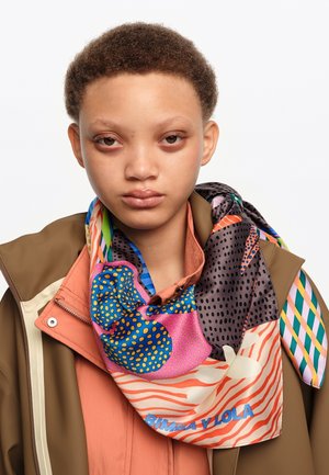 Młoda osoba z krótkimi kręconymi włosami nosi kolorowy szalik w wzory na brzoskwiniowo-zielonej kurtce na jednolitym białym tle.