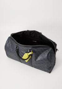 Sac de voyage noir avec un extérieur texturé, deux poignées et une étiquette de bagage jaune. Grande ouverture à zip révélant un intérieur noir.