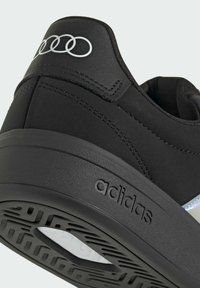 Gros plan sur le talon d'une basket Adidas noire avec logo embossé sur la semelle et anneaux blancs Audi sur la languette arrière.