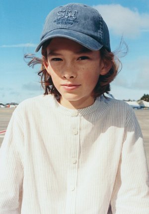 Mlada oseba s svetlimi pegami na obrazu, ki nosi modro kape in svetlo srajco z navpičnimi črtami, stoji na prostem pod delno oblačnim nebom.