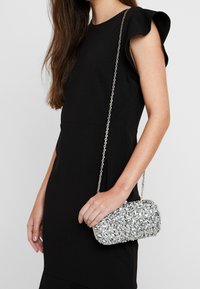 Kobieta w czarnej sukience bez rękawów trzyma małą torebkę z srebrnymi cekinami na cienkim łańcuszku przewieszoną przez ramię.