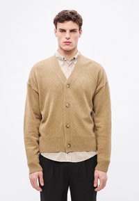 Kamieļkrāsas kardigans no tamborēta auduma ar V veida izgriezumu, pieciem pogas, ribotu apakšmali un iestrādātām piedurknēm, uzvilkts pāri svītrains krekliņš.