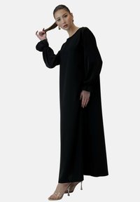 Schwarzes langärmliges Kleid mit gepufften Manschetten, aus glattem Stoff, mit einer lockeren Silhouette. Kombiniert mit transparenten Absatzsandalen.