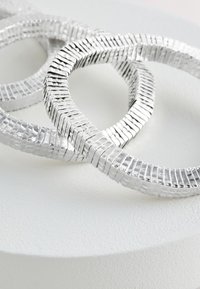 Silver sculptural rings feature a textured, wavy design with a segmented, reflective surface, resting on a textured white pedestal.