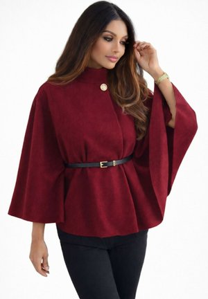 Femme portant un haut ample de style cape bordeaux avec de larges manches, cintré à la taille par une fine ceinture noire, associé à un pantalon noir.
