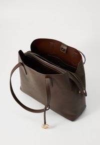 Sac fourre-tout en cuir brun avec deux poignées, poche zippée intérieure, fermeture magnétique et un pendentif logo en ton doré. Texture lisse avec un design minimal.