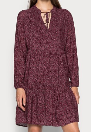 Vrouw draagt een lange mouw, bordeauxroze bloemenjurk met een strikdetail bij de hals en een geplooide taille, staand tegen een eenvoudige achtergrond.