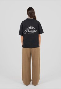 Czarny bawełniany t-shirt z białą grafiką "Zabronione 10119 Berlin" i oliwkowym wzorem liścia z tyłu; połączony z beżowymi spodniami o szerokich nogawkach.