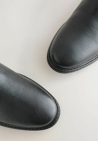 Botas de cuero negro con una textura suave y un diseño de puntera redondeada. Presentan costuras en contraste a lo largo de la suela.