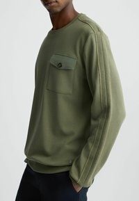Felpa verde oliva con tasca a toppa, dettaglio dei bottoni e strisce laterali testurizzate. Realizzata in materiale morbido con una vestibilità morbida.