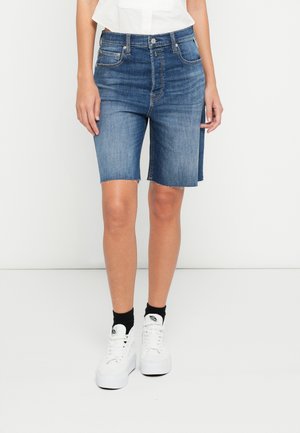 Denimowe bermudy w średnim niebieskim kolorze, z postrzępionym dołem, pięcioma kieszeniami i zapięciem na guzik, zestawione z białymi trampkami i czarnymi skarpetkami.