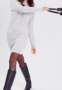 Robe-pull gris clair à maille torsadée avec manches longues et ourlet côtelé, associée à des bottes montantes en cuir noir. Texture lisse et coupe ajustée.