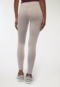 Beżowe legginsy o gładkiej fakturze, z wysokim stanem, zaokrąglonymi szwami z tyłu i w połączeniu z szarymi trampkami.