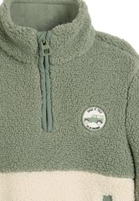 Pull en polaire vert et crème avec zip quart, col montant et patch rond montrant un véhicule tout-terrain vert et du texte sur la poitrine.