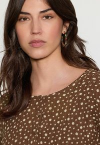 Haut marron à pois avec un col rond, trois boutons au centre et un tissu texturé. Le mannequin porte des boucles d'oreilles colorées.