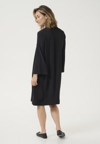 Černé oversized šaty s dlouhými rukávy, hladkou texturou, ke kolenům sahajícím hemem a jednoduchým designem. Nosené s černými plochými botami.