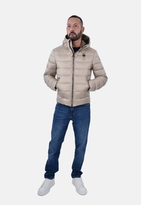 Giubbotto imbottito beige realizzato in materiale liscio e imbottito, con cappuccio, zip frontale e dettaglio del logo; abbinato a jeans blu e sneaker bianche.