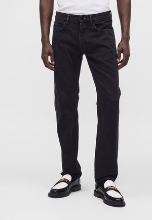 Homme portant un jean noir droit avec des mocassins blancs et noirs et une chemise blanche, debout devant un fond uni.
