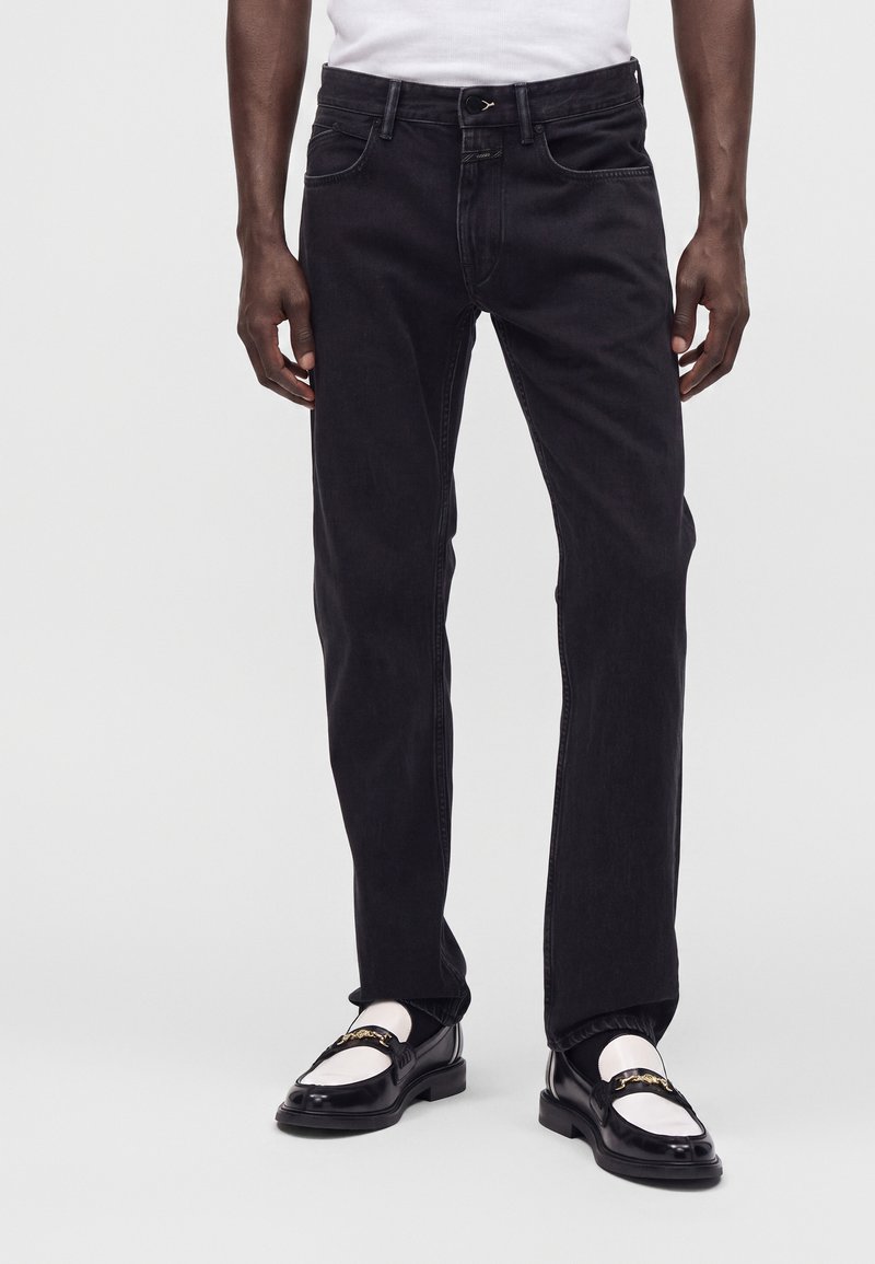 Homme portant un jean noir droit avec des mocassins blancs et noirs et une chemise blanche, debout devant un fond uni.