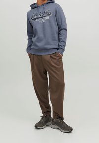 Szary hoodie z białym tekstem logo, w zestawie z luźnymi, brązowymi spodniami i ciemnoszarymi trampkami. Swobodny, wygodny zestaw ubrań.