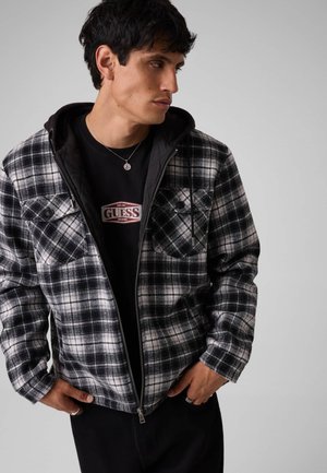Man draagt een zwart-wit geruite jas met ritssluiting over een zwart Guess T-shirt, kijkt naar links met handen in de zakken.