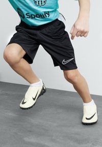 Svarta sportshorts med sidostråkar, tillsammans med gul-vita Nike fotbollsneakers med svarta accenter och en texturerad sula, på grå yta.