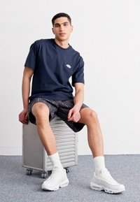 Marineblå bomulds t-shirt med et lille logo, kombineret med stribede shorts og hvide sneakers. Model siddende på en sølvmetalbeholder.