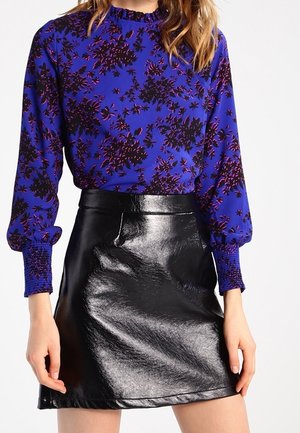 Kvinde iført en blå langærmet blomstret bluse med rynkede manchetter og en sort skinnende lædermininederdel med høj talje.