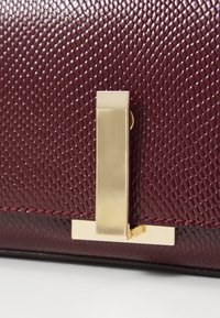 Borsa a mano in pelle textrurizzata borgogna con chiusura e finiture dorate, dotata di una base liscia a contrasto e dettagli sottili.