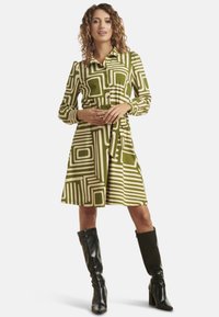 Groene jurk met een patroon van witte geometrische ontwerpen, knoopsluiting aan de voorkant, lange mouwen, riem op de taille, gecombineerd met zwarte kniehoge laarzen.