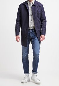 Navy blue lightweight trench coat with a collared neckline, button closure, worn over a light shirt and blue jeans, paired with white sneakers.