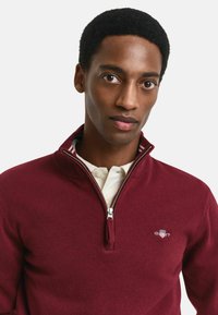 Burgunderfarbener Halbzip-Pullover aus weichem Strickmaterial, mit einem silbernen Reißverschluss und einem runden Logo auf der Brust.