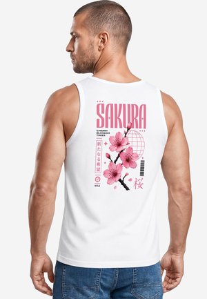 Mann trägt weißes Tanktop mit pinkem Kirschblütenzweig und Text "Sakura" auf dem Rücken, steht vor einem einfarbigen Hintergrund.